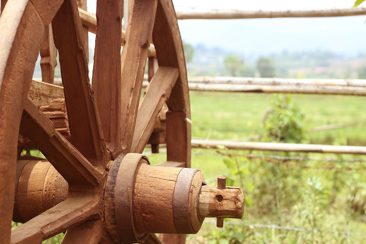 Une structure de roue en bois, probablement un élément d’une charrette ou d’une machine, avec un paysage rural en arrière-plan.