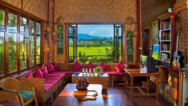 Ein Zimmer mit einem großen Fenster und Blick auf eine grüne Landschaft, ein Sofa mit roten Kissen, ein Tisch mit Teekanne und ein Bücherregal.