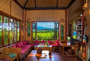 A room with a large window offering a view of a green landscape, a sofa with red cushions, a table with a teapot, and a bookshelf. - Mr. Charles Riverview Lodge (Hsipaw)