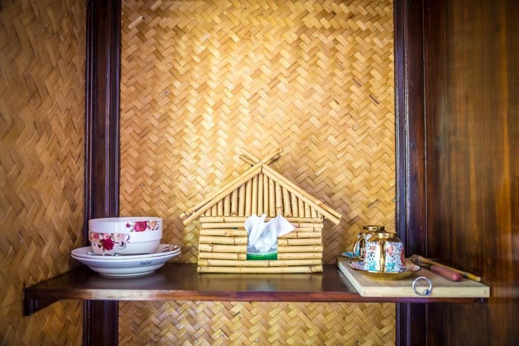 Une pièce avec un mur en bambou tressé, une étagère en bois sur laquelle repose un bol décoré de motifs floraux, une petite structure ressemblant à une maison et un pot décoratif.