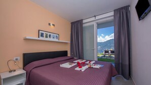 A hotel room with a bed, a bedside table, a TV, a window with curtains, and a view of the outdoors. - Casa Laura Apt. 3, Plesio, Italy (Plesio)