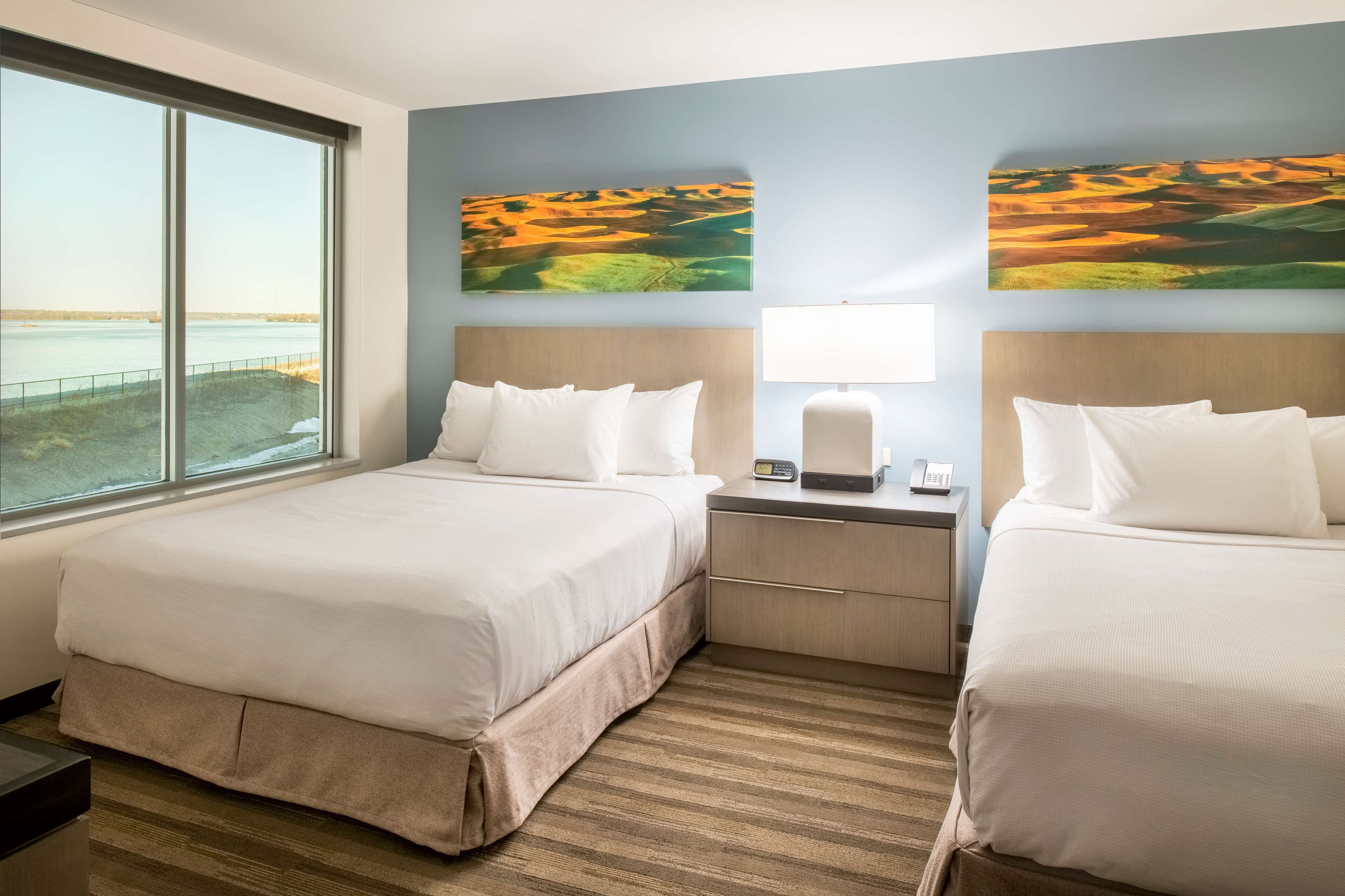 A hotel room with two beds, a nightstand, a lamp, and a large window with a view of the beach.