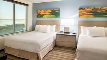 A hotel room with two beds, a nightstand, a lamp, and a large window with a view of the beach.