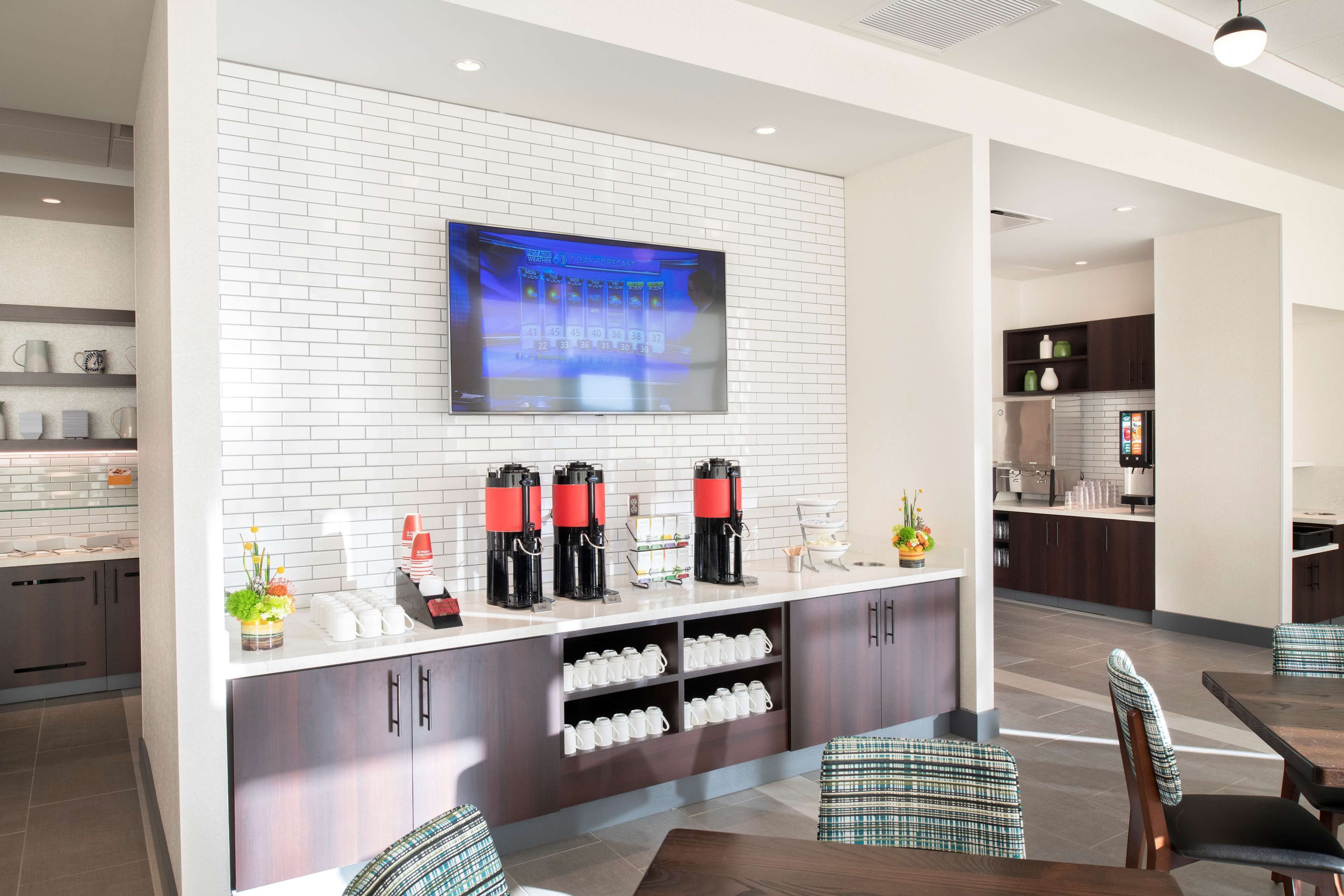 A modern kitchen with a flat-screen TV mounted on a white brick wall, a coffee station with multiple coffee makers, and a dining area with checkered upholstered chairs.