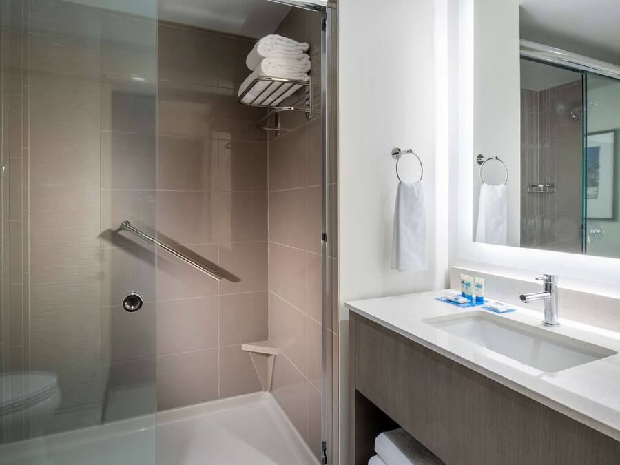 Une salle de bain moderne avec une douche à parois de verre, un lavabo blanc avec un miroir et des serviettes soigneusement pliées sur une étagère.