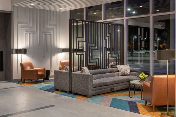 Un moderno vestíbulo de hotel con un sofá seccional, dos sillones, una mesita con frutero y un separador decorativo.