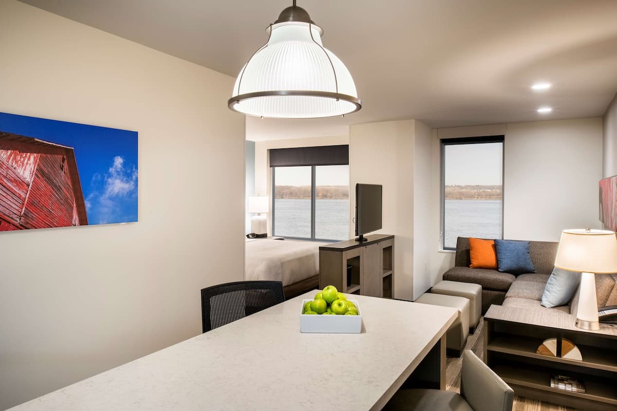 Une chambre d’hôtel moderne équipée d’une table à manger, d’un canapé, d’une tél évision et d’une grande fenêtre donnant sur l’eau.