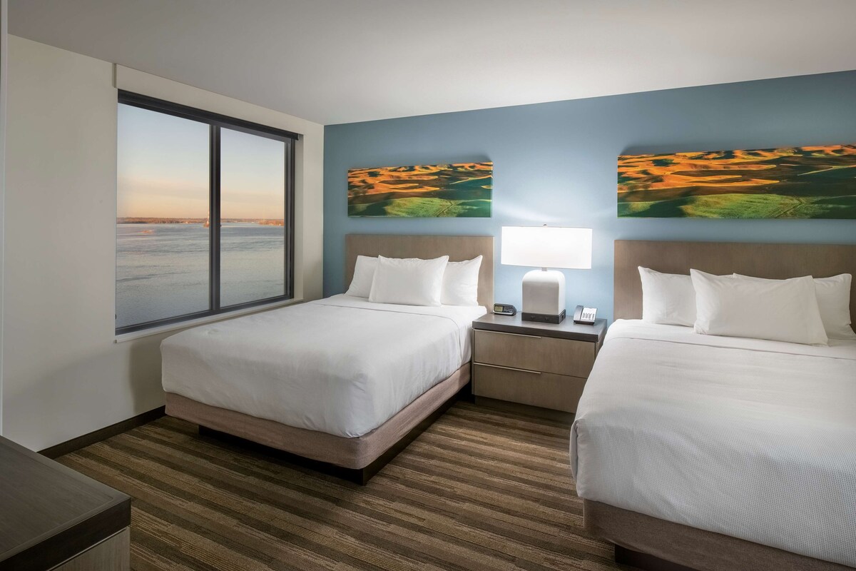 Habitación de hotel con dos camas, una mesita de noche con lámpara, una ventana con vista al mar y dos cuadros de paisajes en la pared.