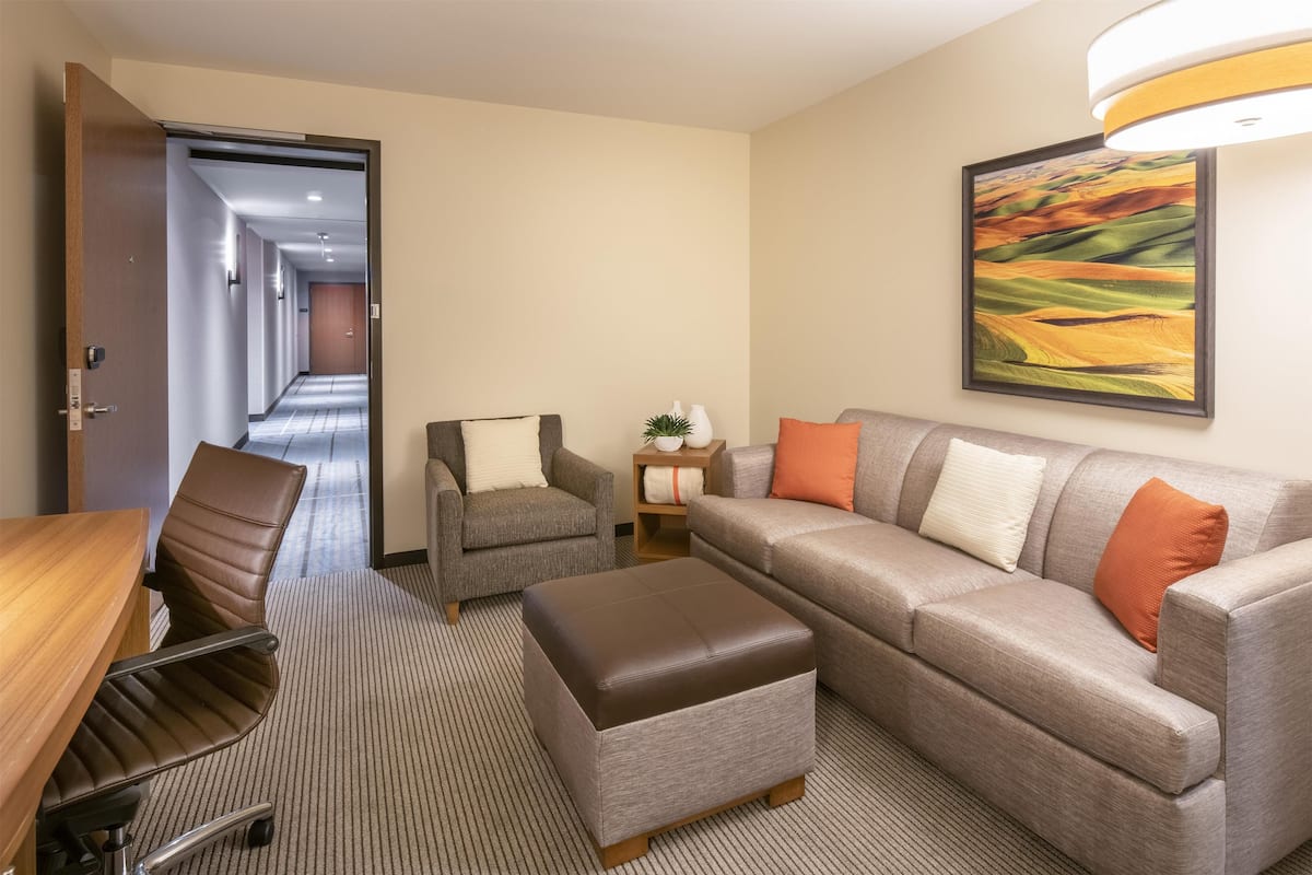 Ein Hotelzimmer mit Sofa, Ohrensessel, Hocker, Schreibtisch und Lampe.