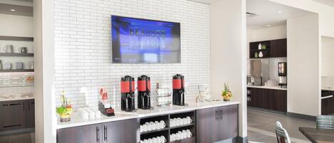 A modern kitchen with a flat-screen TV mounted on a white brick wall, coffee machines, and a counter with mugs.