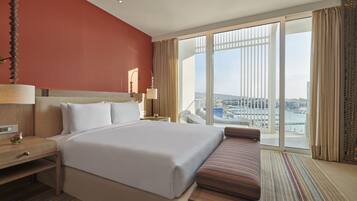 A hotel room with a large bed, a bench, a nightstand, a lamp, and a view of a cityscape through large windows.