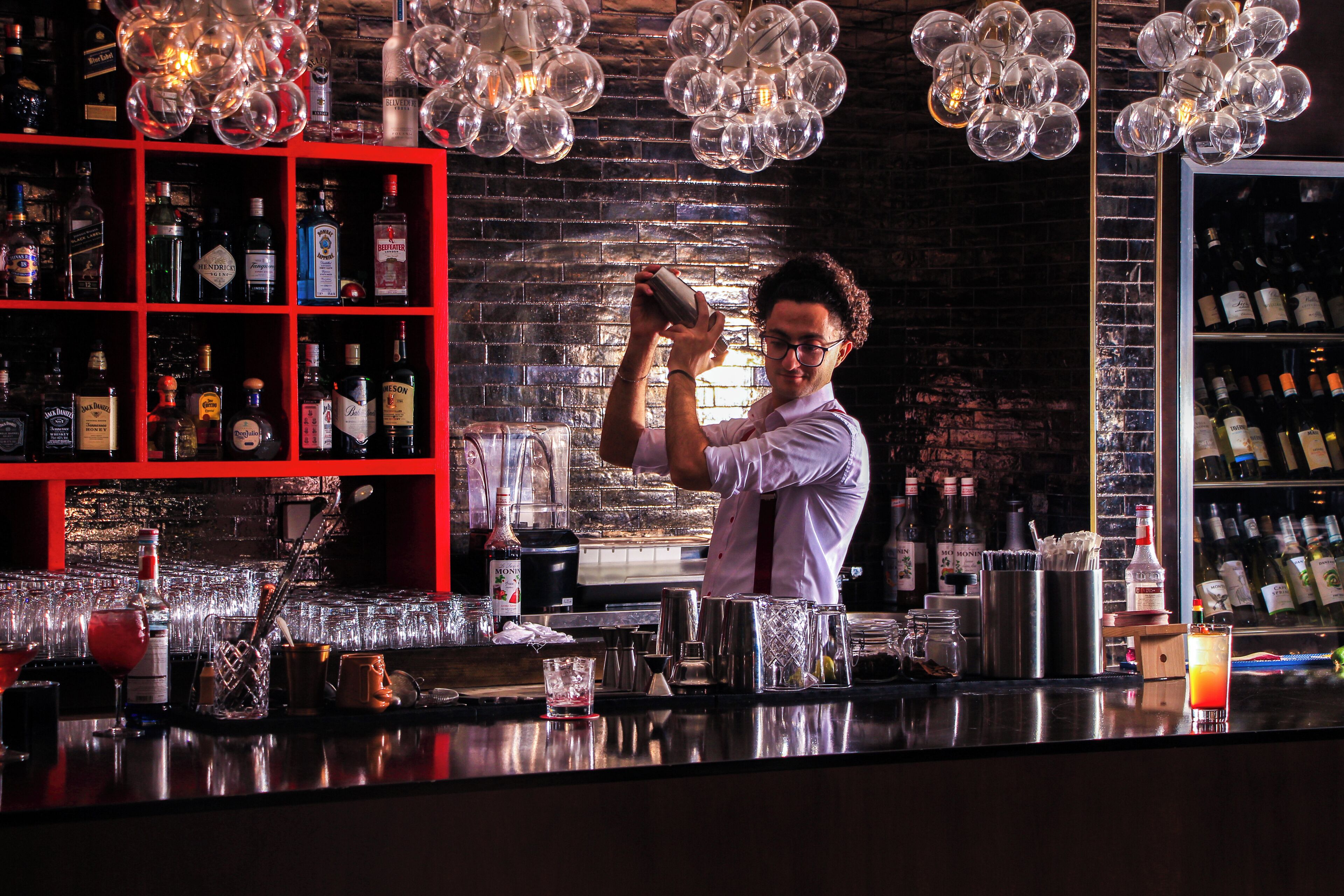 A bartender preparing a drink at a bar with a brick wall background and shelves of liquor bottles.