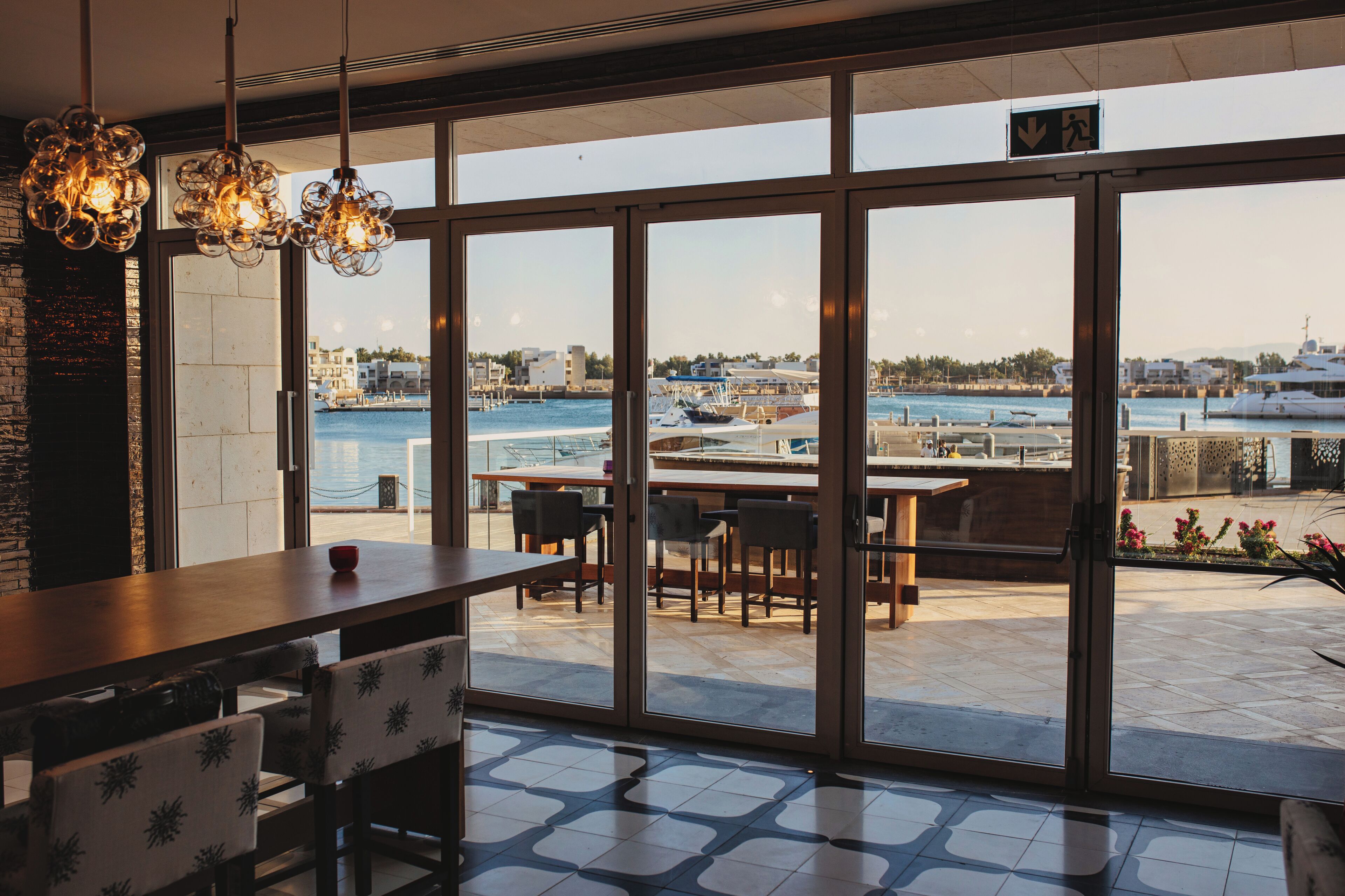 A restaurant with large windows overlooking a marina, featuring a wooden table and chairs, and hanging pendant lights.