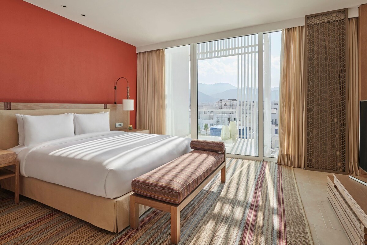 A hotel room with a large bed, a bench, a TV, and a view of a cityscape through large windows.