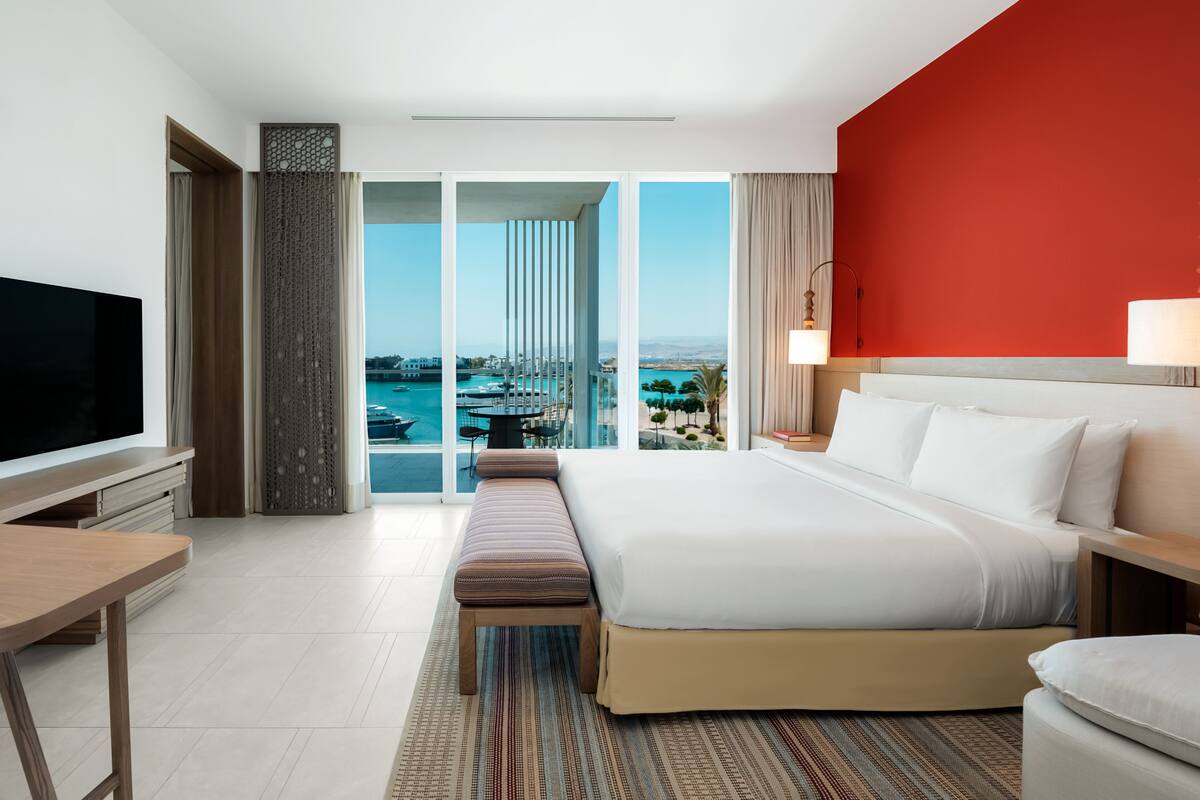 A modern hotel room with a large bed, a TV, and a balcony with a view of the city and water.