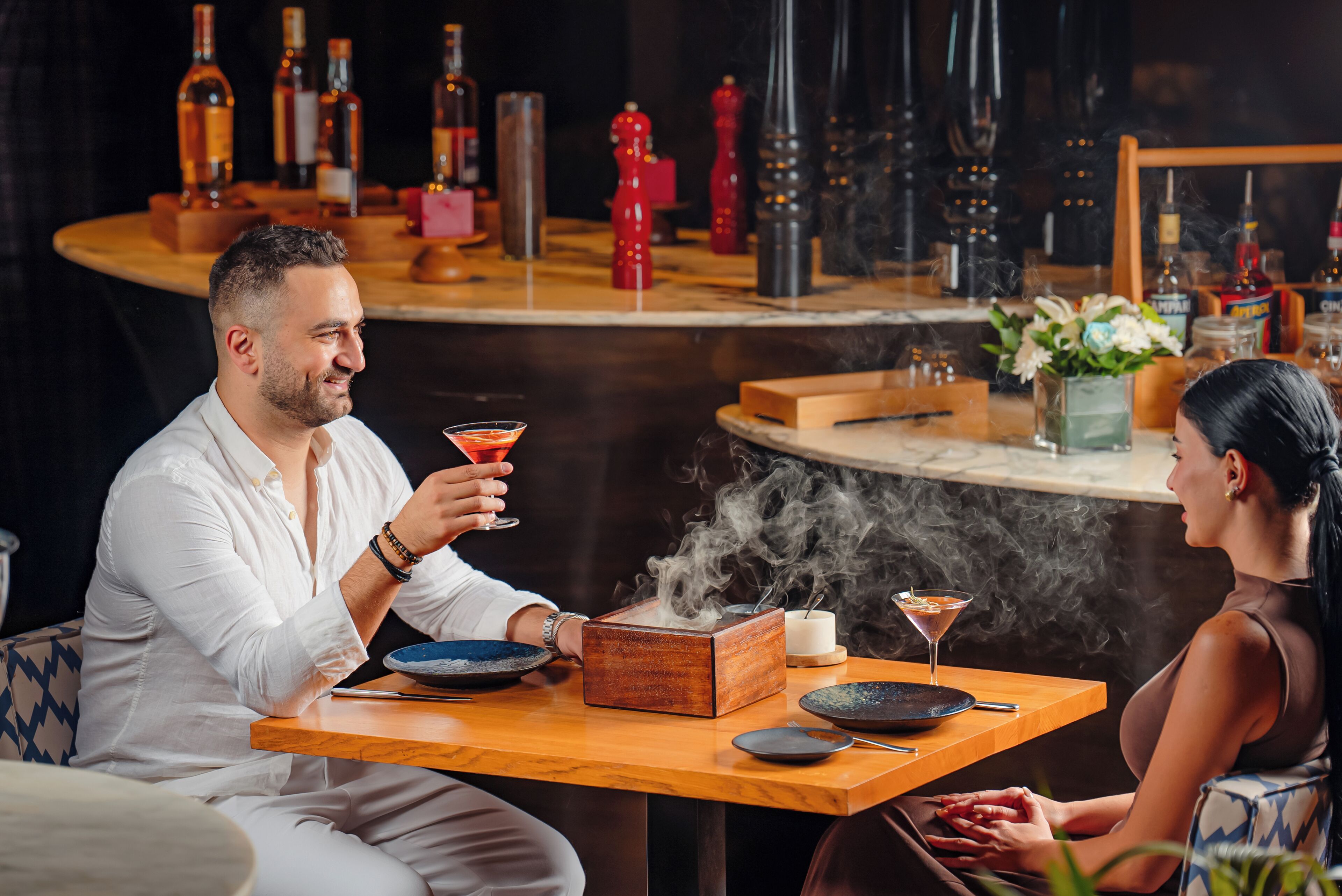 Un couple est assis à une table en bois, avec un comptoir de bar en arrière-plan, où l’on peut voir différentes bouteilles et une bougie.