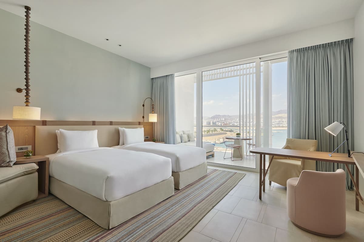 A hotel room with two beds, a desk, a chair, a view of the city, and a large window with curtains.