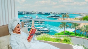 A person in a white bathrobe reading a book on a balcony with a view of a marina and boats.
