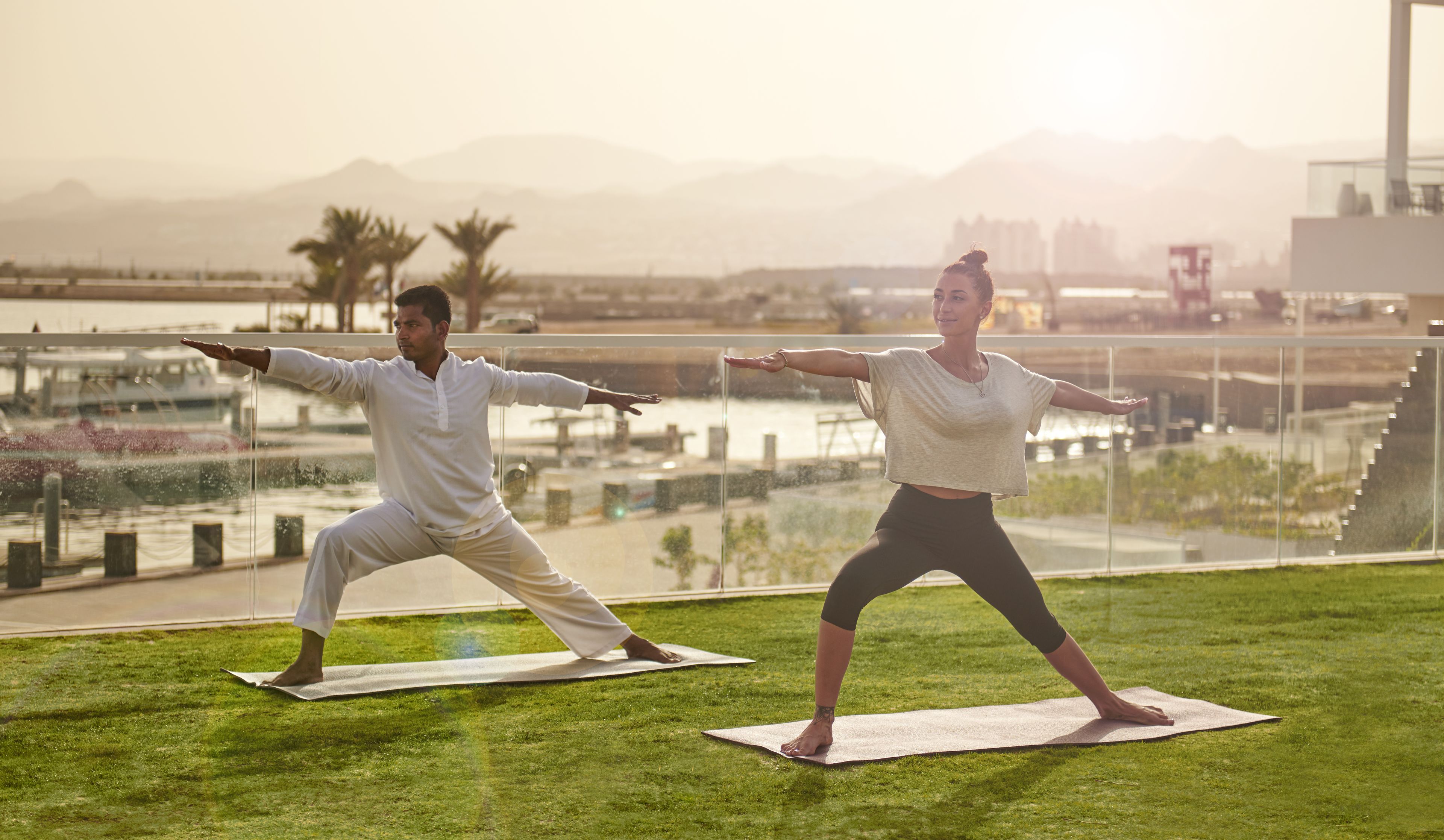 Two people practicing yoga on a grassy rooftop with a cityscape view in the background.