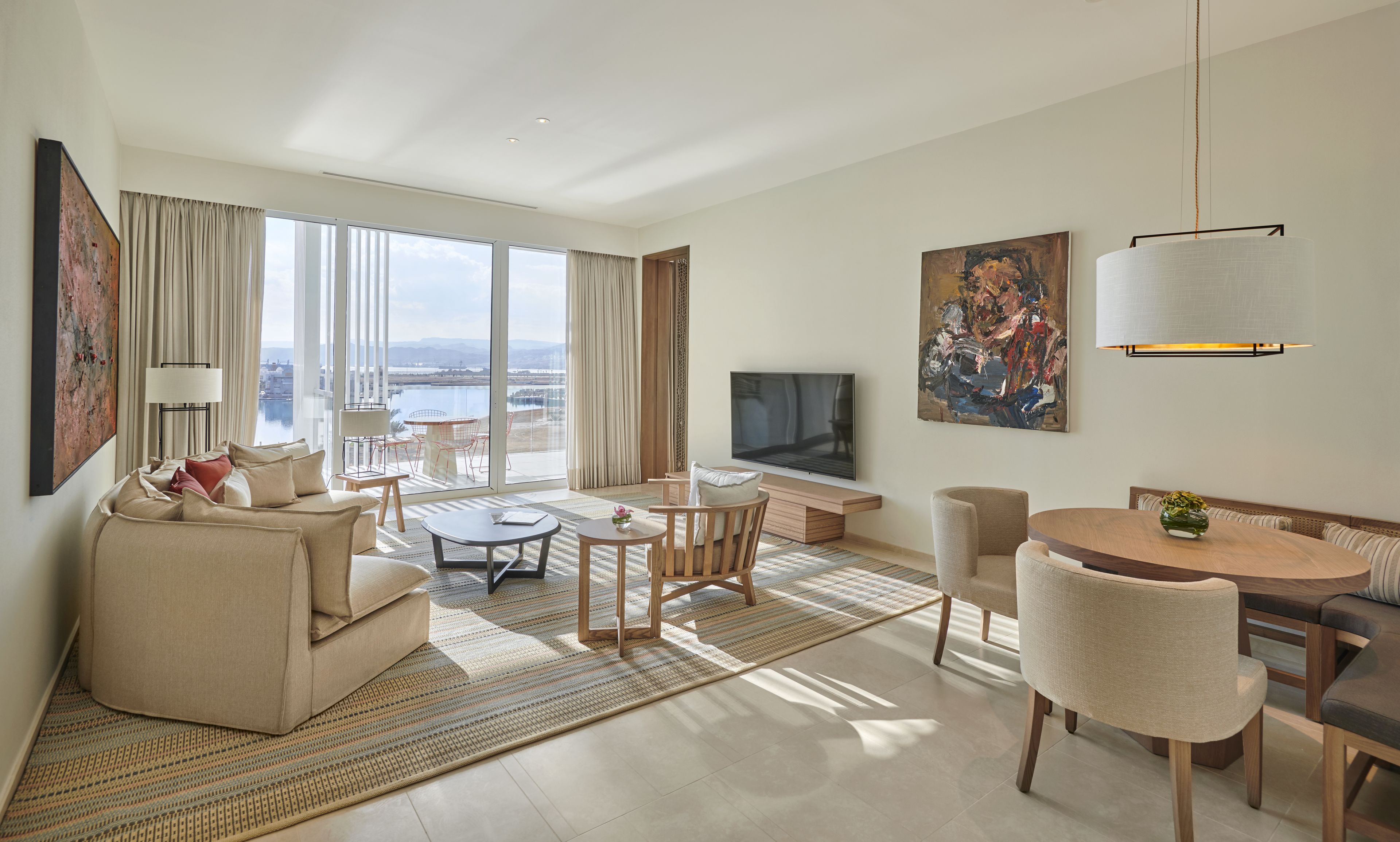 A modern hotel room with a large window, a flat-screen TV, a dining area, and a sofa.