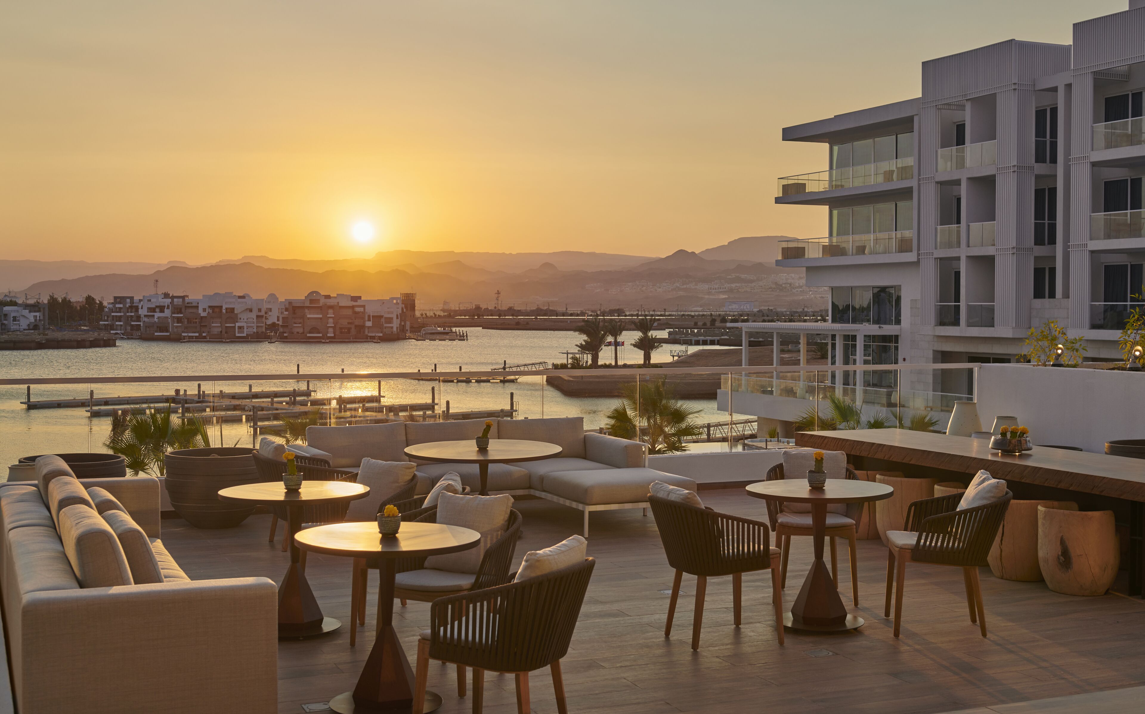 Une terrasse sur le toit avec des sièges extérieurs, une vue sur le coucher de soleil au-dessus d’une étendue d’eau et des bâtiments modernes en arrière-plan.
