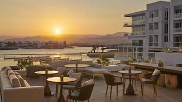 Terraço no telhado com área de estar ao ar livre, vista do pôr do sol sobre a água e edifícios modernos ao fundo.