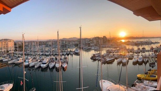 Un puerto deportivo con barcos amarrados, un atardecer de fondo y un edificio con balcones.