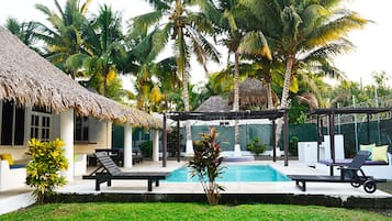 A pool area with lounge chairs, a thatched-roof structure, and palm trees.