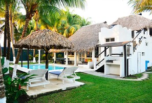 Un espace piscine comprenant deux chaises longues, un parasol en chaume et un bâtiment blanc avec un escalier.