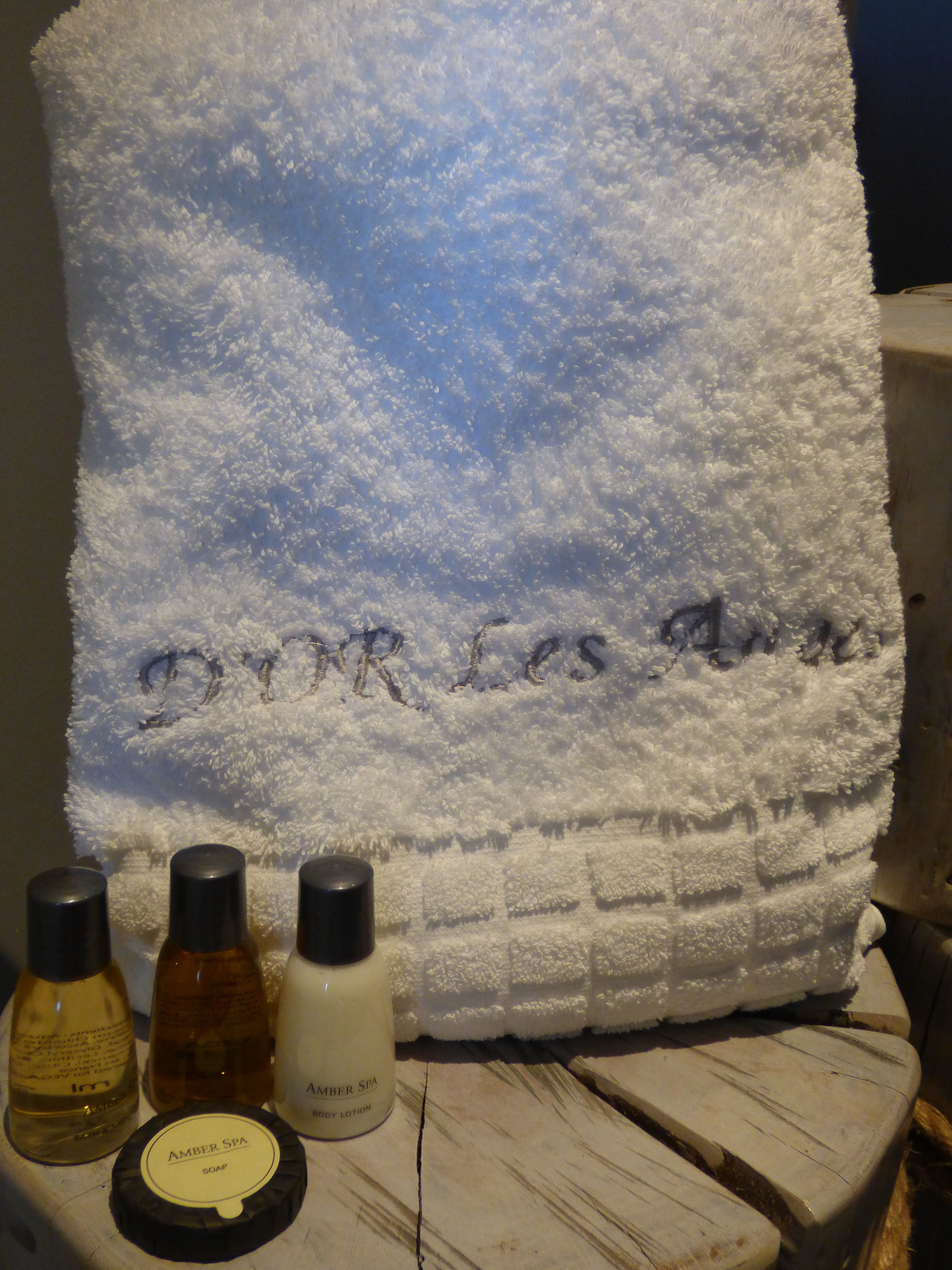 A white towel with "DOR LES AME" embroidered on it, placed on a wooden surface with three bottles of soap and lotion in front.