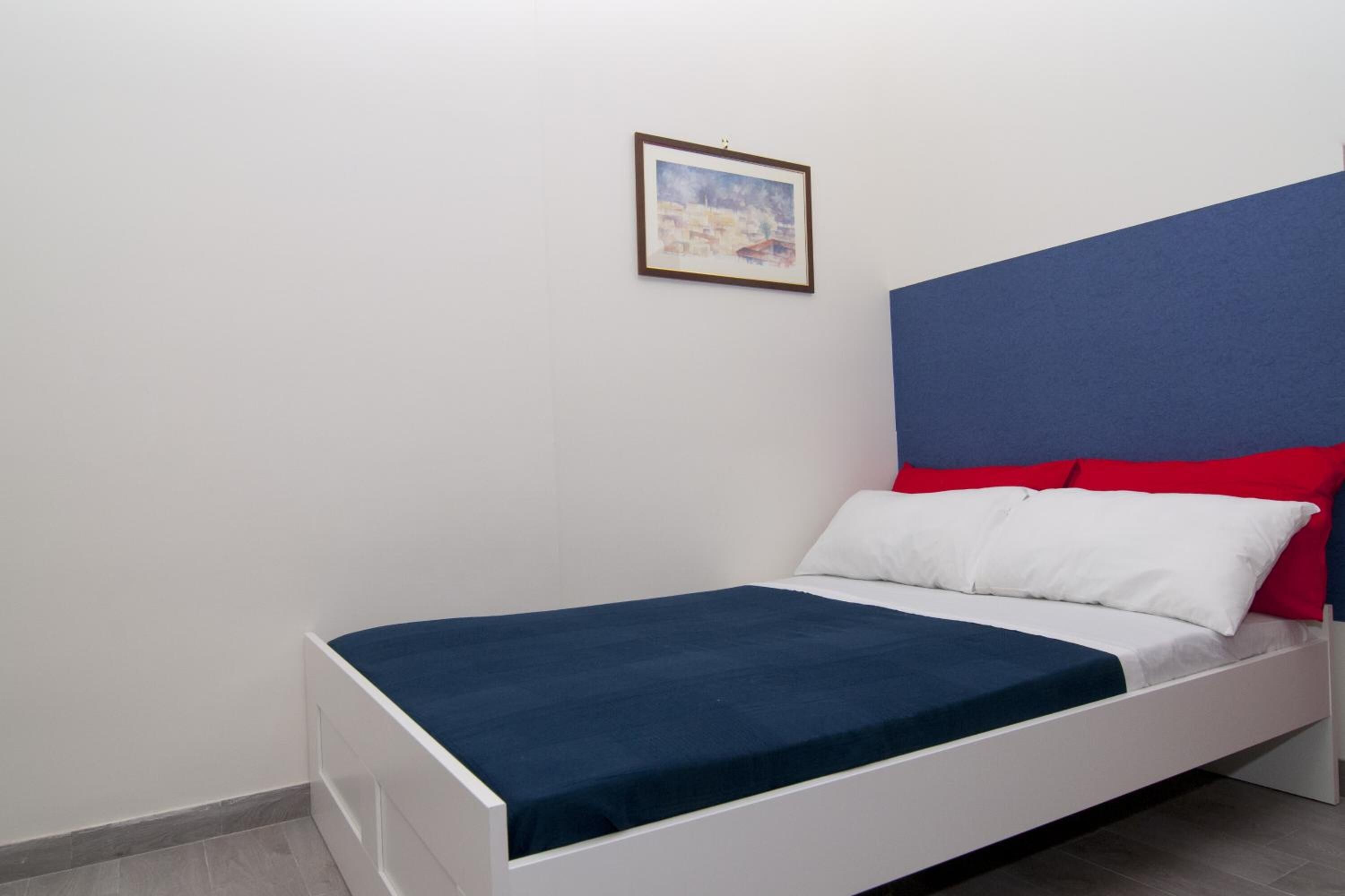 Una cama individual con estructura blanca, cubierta de colchón azul y almohadas rojas y blancas. Encima de la cama, hay un cuadro en la pared.