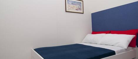 Una cama individual con estructura blanca, cubierta de colchón azul y almohadas rojas y blancas. Encima de la cama, hay un cuadro en la pared.
