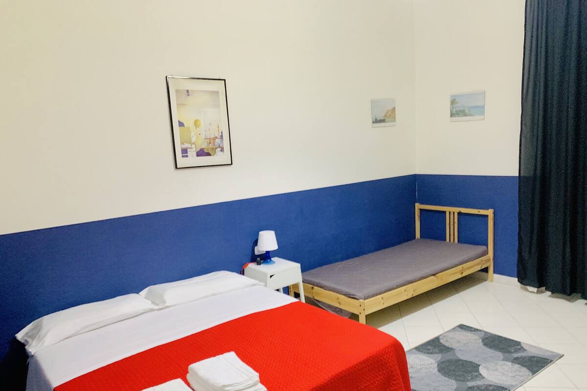 A small, single-room hotel room with a bed, a nightstand, a lamp, a small table, and a window with curtains.