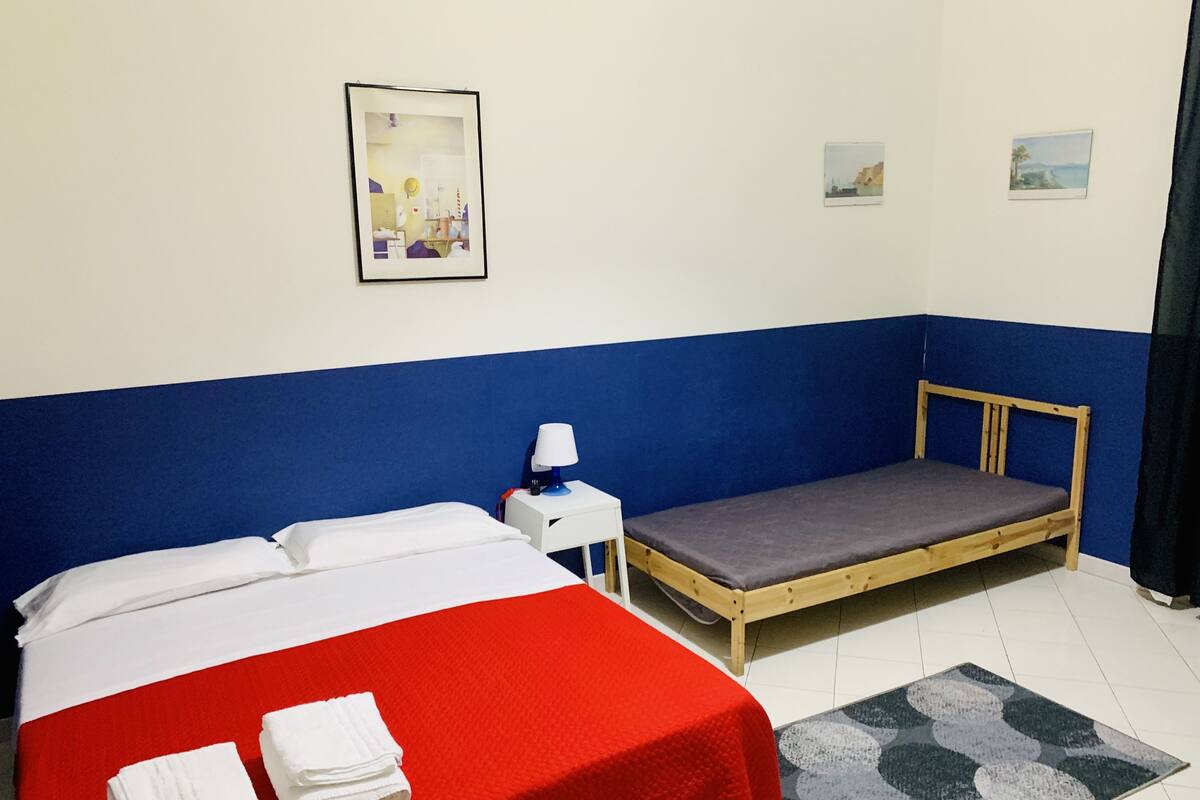 A room with a bed, a small bedside table, a nightstand, a single bed, a red bedspread, a grey mattress, a blue wall, a white wall, a framed picture, and a small rug.