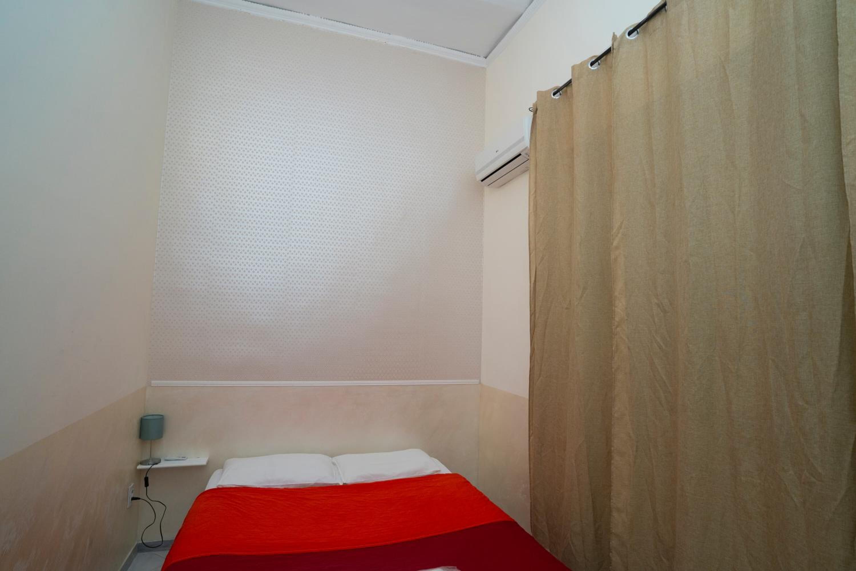Una habitación pequeña con una cama individual, una colcha roja, un almohadón blanco, una mesita de noche con una lámpara y una cortina color beige.