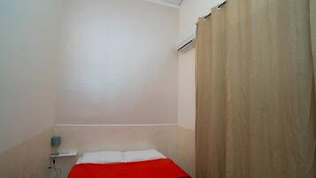 Una habitación pequeña con una cama individual, una colcha roja, un almohadón blanco, una mesita de noche con una lámpara y una cortina color beige.