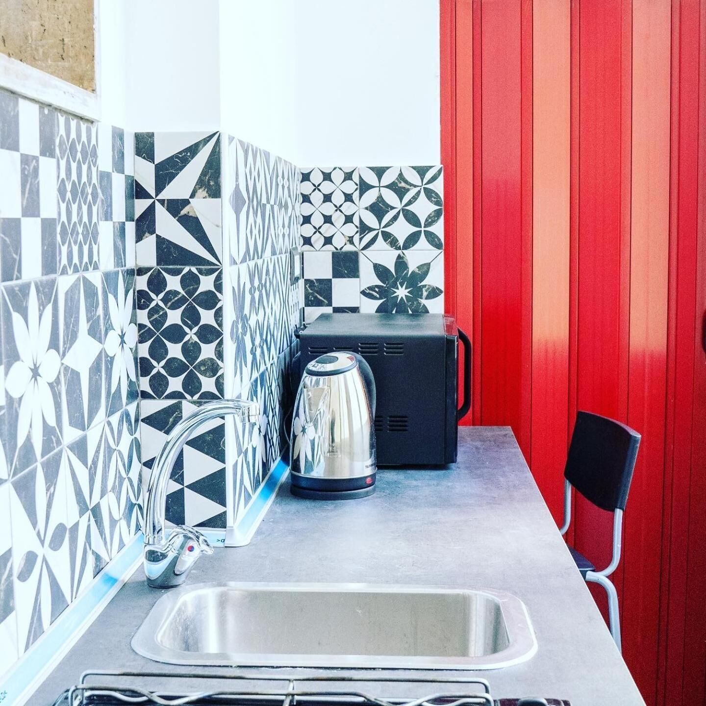 A compact kitchen with a sink, kettle, and toaster. The walls are tiled with geometric patterns.