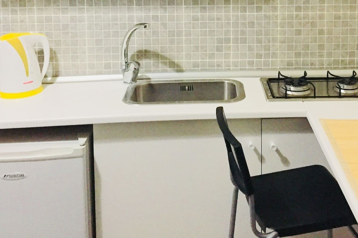 A compact kitchen with a sink, two gas stoves, a kettle, and a small refrigerator.