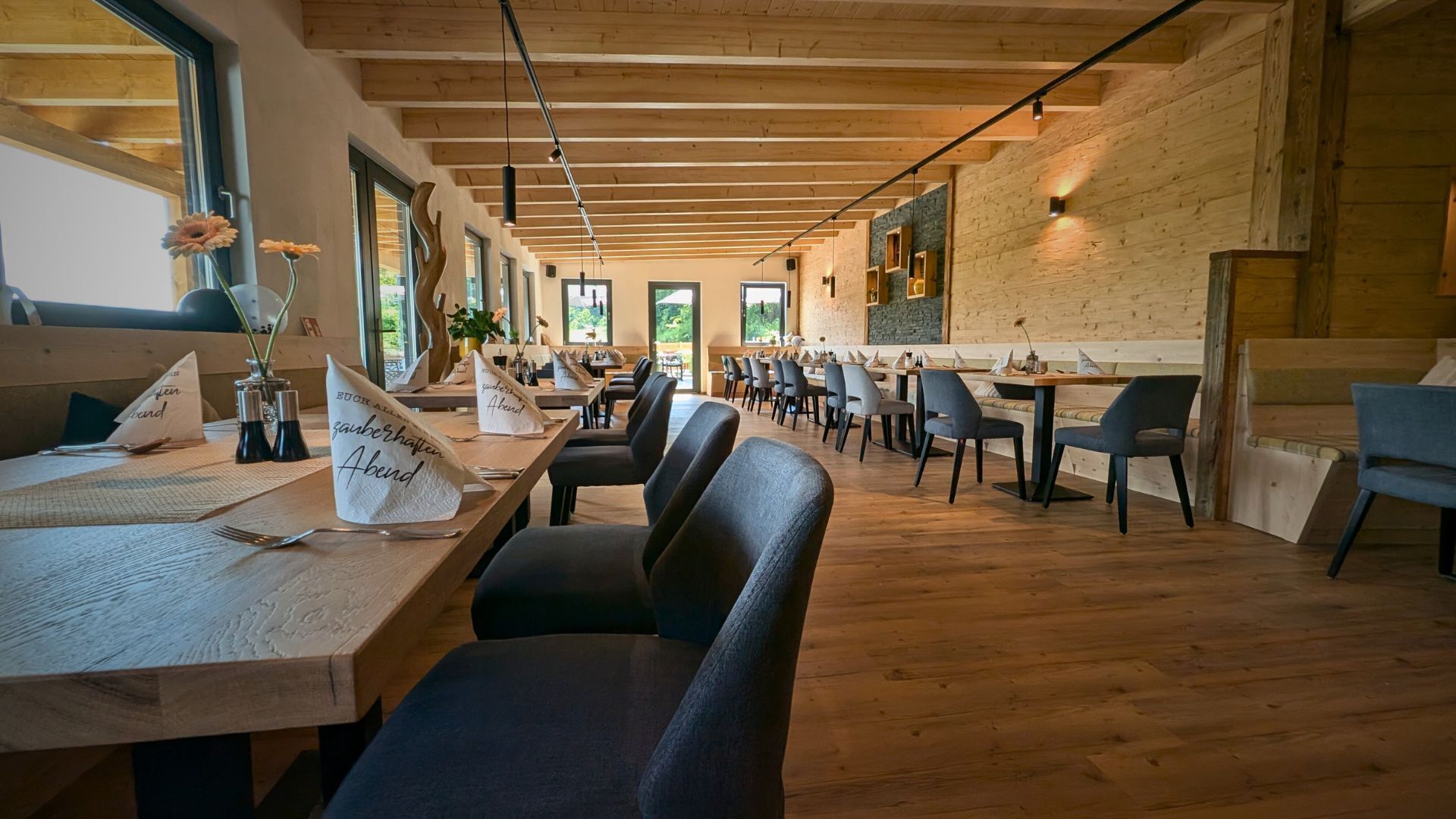 A restaurant with wooden tables and chairs, large windows, and a high ceiling.