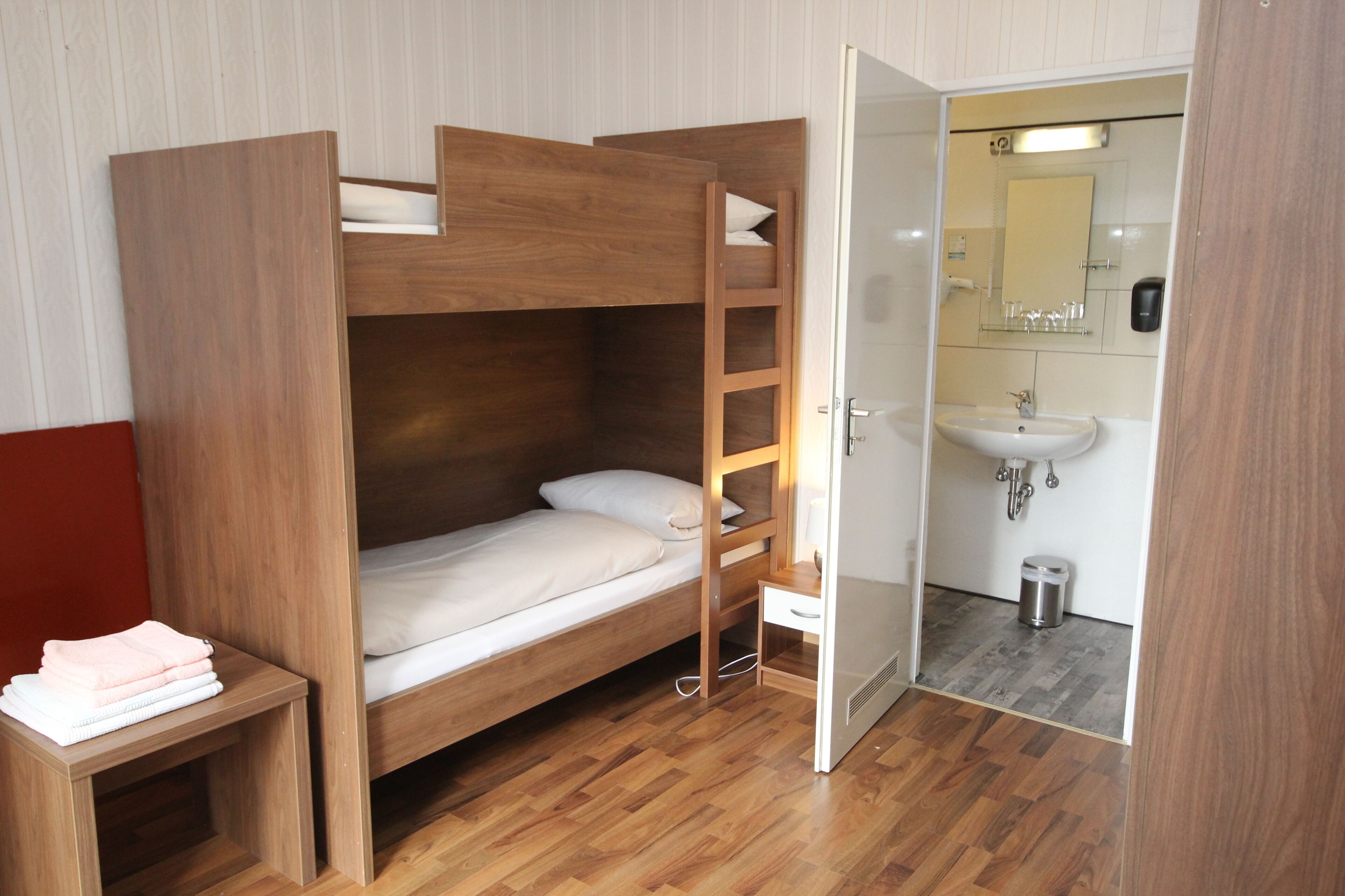 A bunk bed with a wooden frame, a small bedside table with towels, and a bathroom with a sink and mirror.