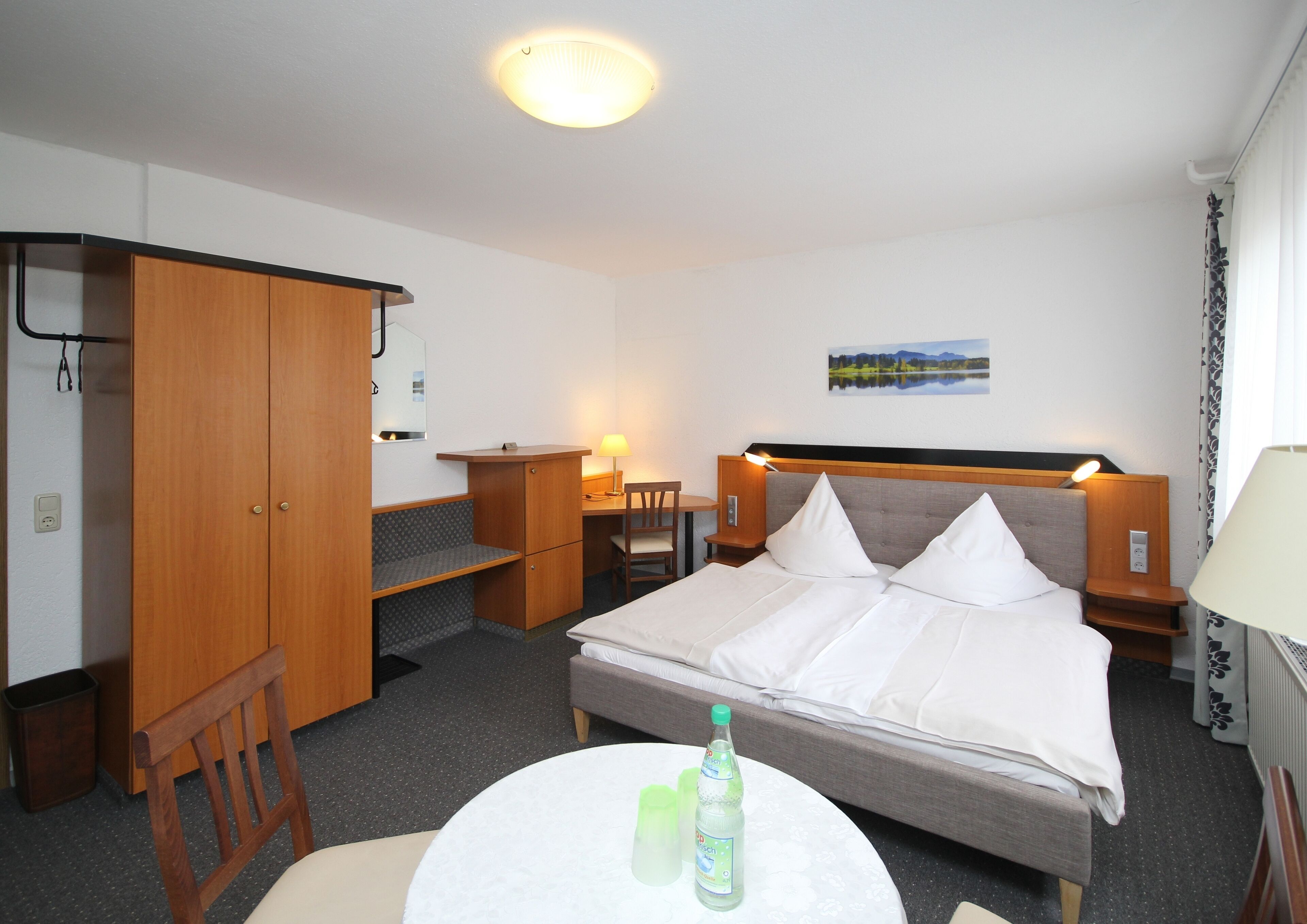 A hotel room with a bed, a desk, a chair, a wardrobe, and a small table with a bottle.