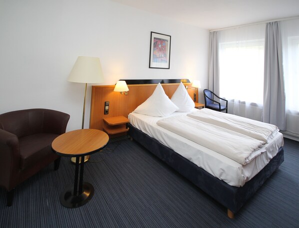 A hotel room with a bed, a chair, a small table, and a nightstand with a lamp.