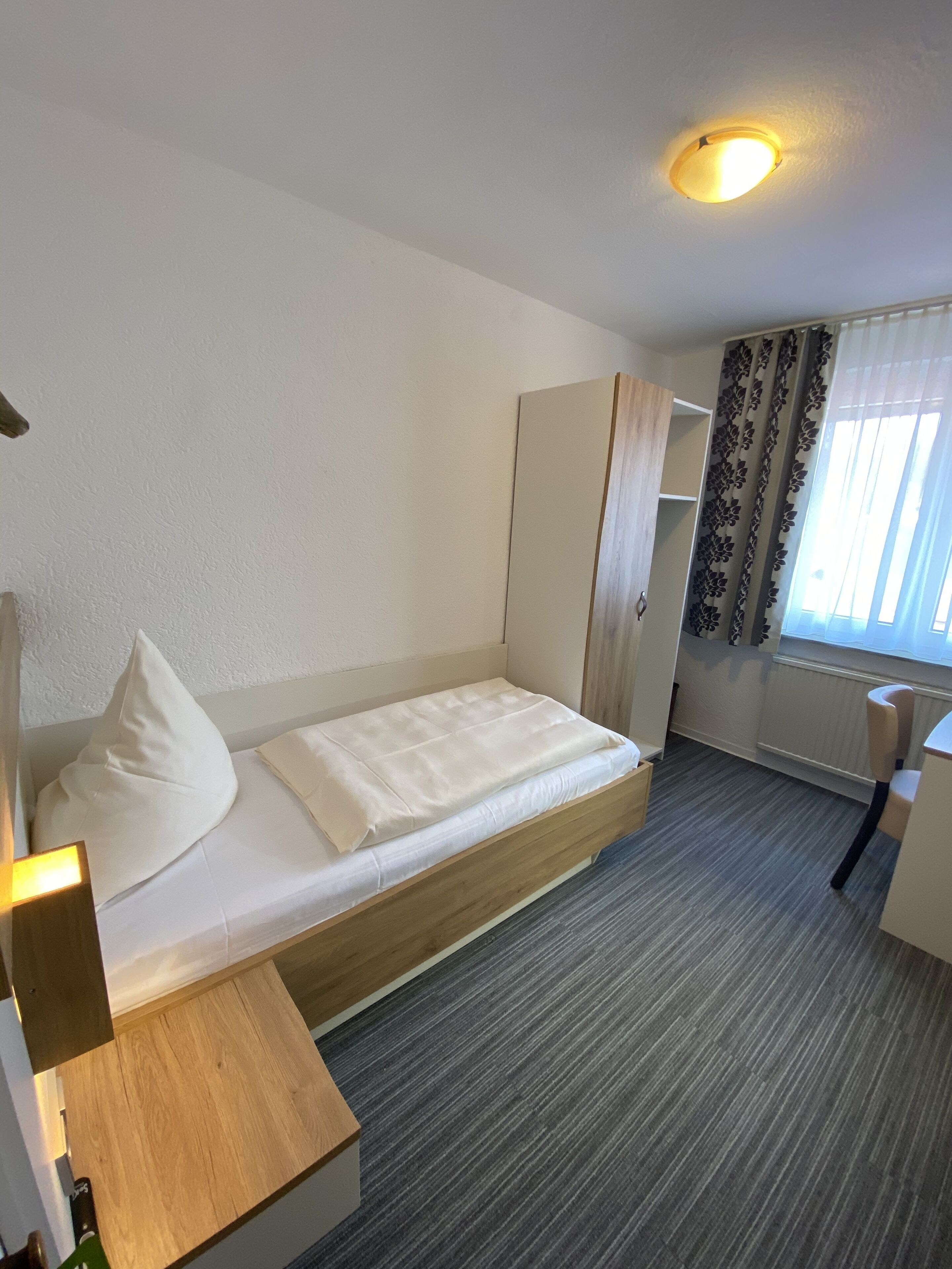 A single-bed room with a wooden headboard, a bedside table, a wardrobe, and a window with curtains.