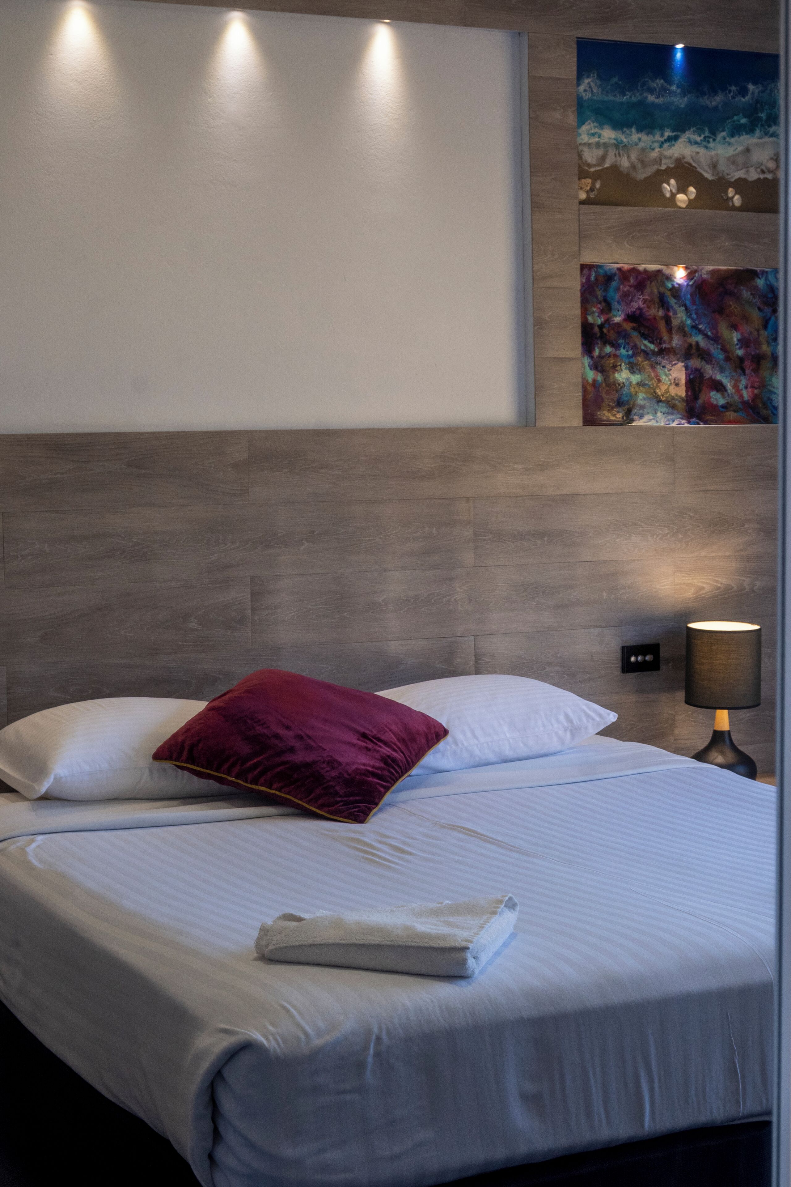 Un lit bien fait, avec des draps blancs et un seul oreiller rouge, une lampe de chevet et une œuvre d’art accrochée au mur représentant une scène de plage.