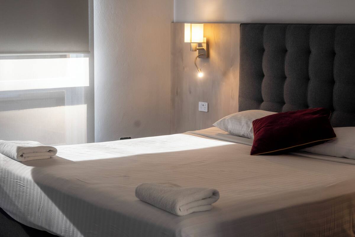 Una habitació d'hotel amb un llit ben fet, una làmpada penjada a la paret i una finestra amb persianes.
