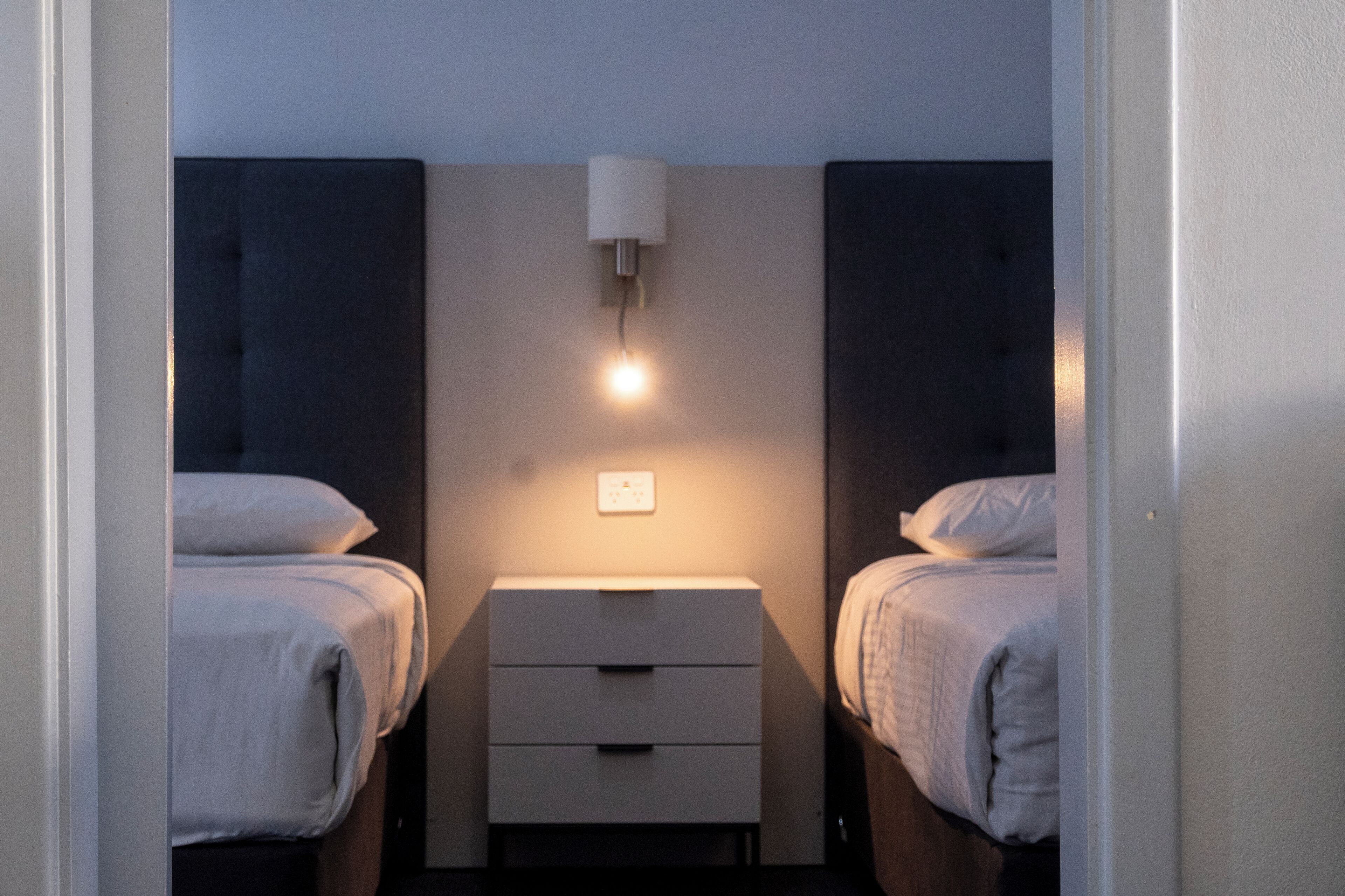 A small, modern bedroom with two beds, a bedside table, and a wall-mounted light fixture.