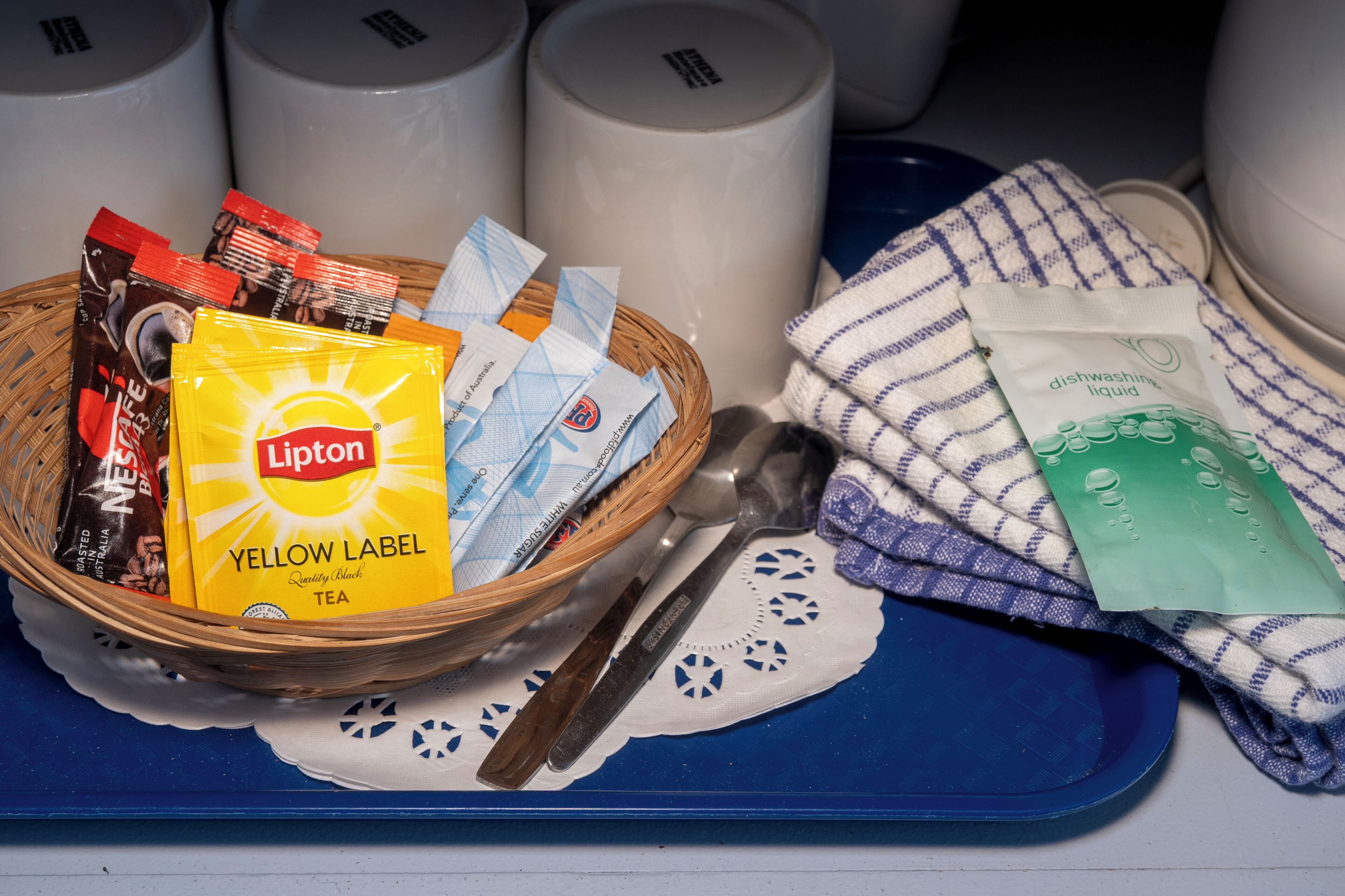 A basket with Lipton tea and coffee packets, a spoon, and a dishwashing liquid packet on a blue tray with a white doily.
