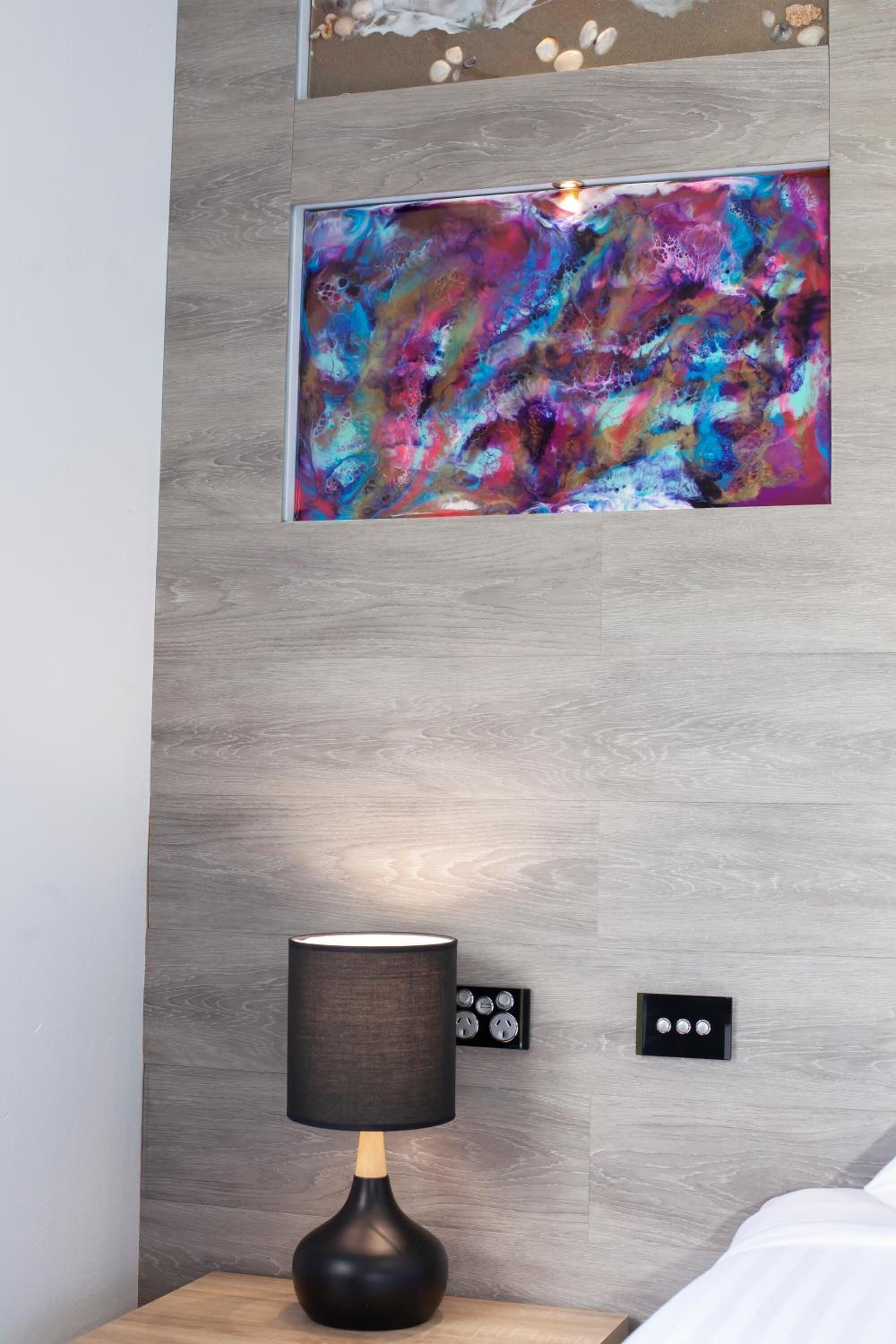 A modern bedroom with a wooden headboard, a bedside table with a lamp, and a colorful abstract painting on the wall.