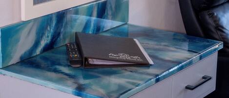 A room with a glass-top desk, a remote control, a notebook, a mirror, a television, and a black leather chair.