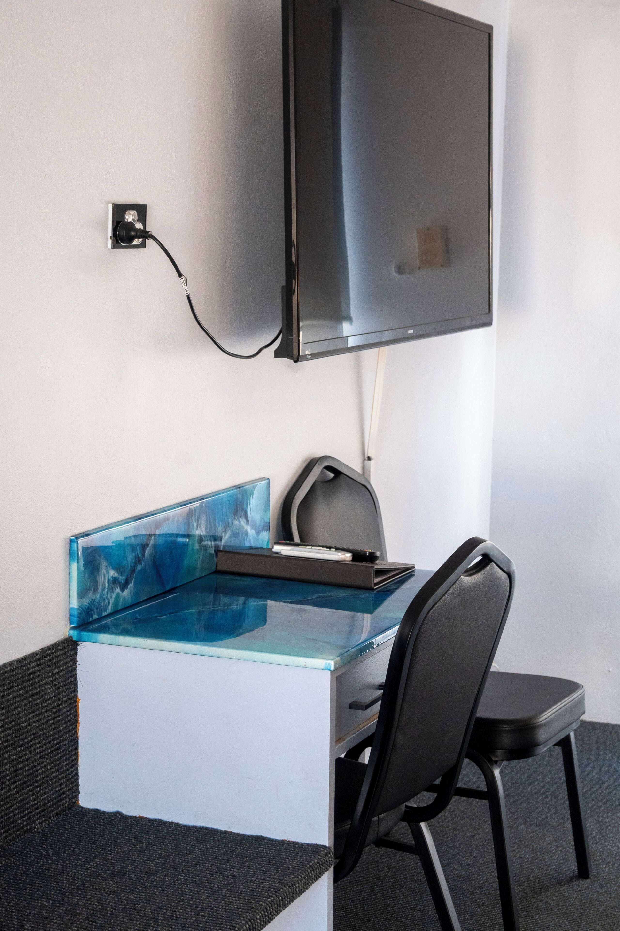A modern office desk with a glass top, a black chair, a flat-screen monitor, and a power outlet on the wall.